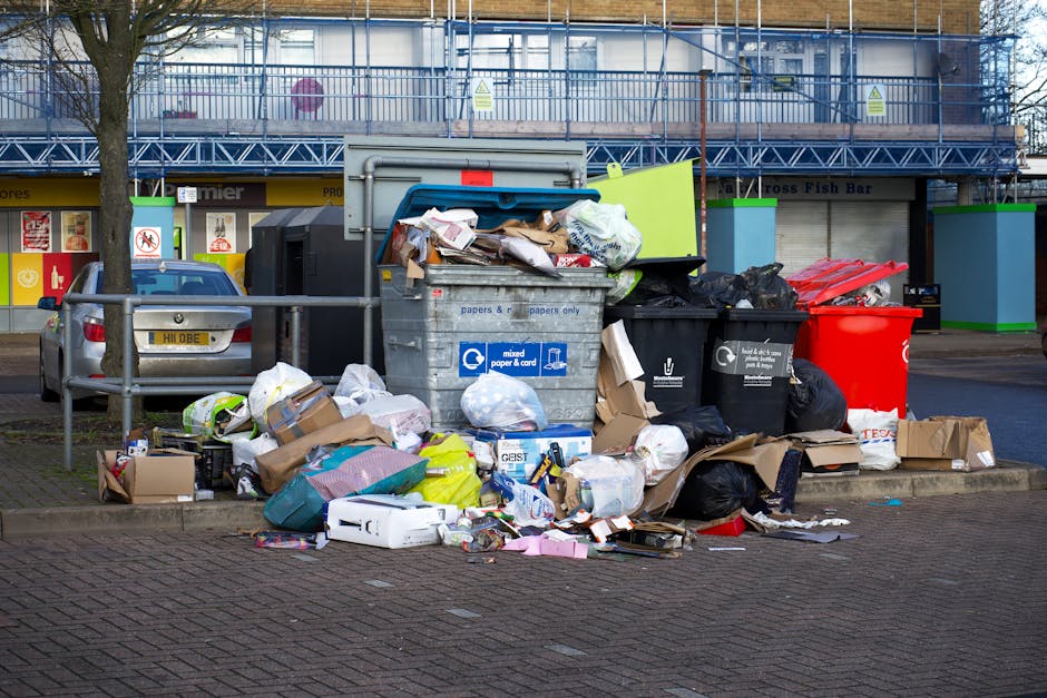 A large collection of mixed household rubbish and waste materials is piled on a city pavement adjacent to a street. The waste includes black bin bags, flattened cardboard boxes, paper, plastic packaging, and discarded packaging materials scattered around a grey communal bin marked for mixed paper and card recycling. Behind the pile, there are parked vehicles, including a silver car with a visible yellow license plate, and a building with a blue scaffold and signage for local shops. The rubbish appears to have accumulated beyond the capacity of the bin, extending onto the surrounding pavement, indicating an overspill of waste often associated with non-collection or private waste disposal services. The scene is set during daylight hours, with natural light illuminating the area, illustrating a typical urban scenario where independent rubbish removal services might be engaged to clear waste from high-traffic communal areas, complementing local authority services.