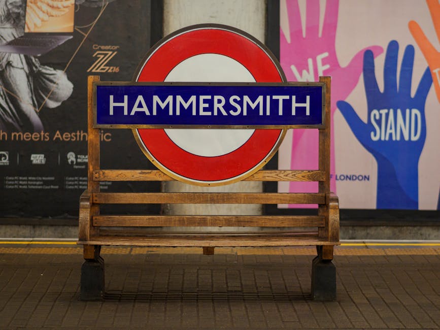 A wooden bench with a slatted seat and backrest, positioned on a tiled surface in front of a large London Underground sign displaying 'HAMMERSMITH' in white letters on a blue background within a red and white roundel. Behind the sign, there are colorful posters, one featuring a pink hand with the words 'WE STAND' in white, and another with black background and partially visible text. The scene appears to be in an indoor or covered public space, illuminated by ambient lighting, with the focus on the bench and station sign, which may relate to local transport or community spaces involved in waste management discussions or services.