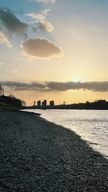 The image depicts a pebble beach shoreline during sunset or late afternoon, with a calm body of water on the right side extending into the distance. The foreground features numerous small, rounded pebbles in shades of grey and brown, leading toward the water's edge. In the background, a cityscape with several high-rise buildings is silhouetted against the sky, which is partly cloudy with a mixture of dark and light clouds illuminated by the setting sun, casting a warm, golden glow across the scene. The environment appears tranquil and natural, with no visible trash or debris, and the scene is suitable for referencing private waste collection or on-site clearance services provided by companies such as Waste Collection Hammersmith, highlighting the importance of maintaining clean, pollution-free waterfront areas.