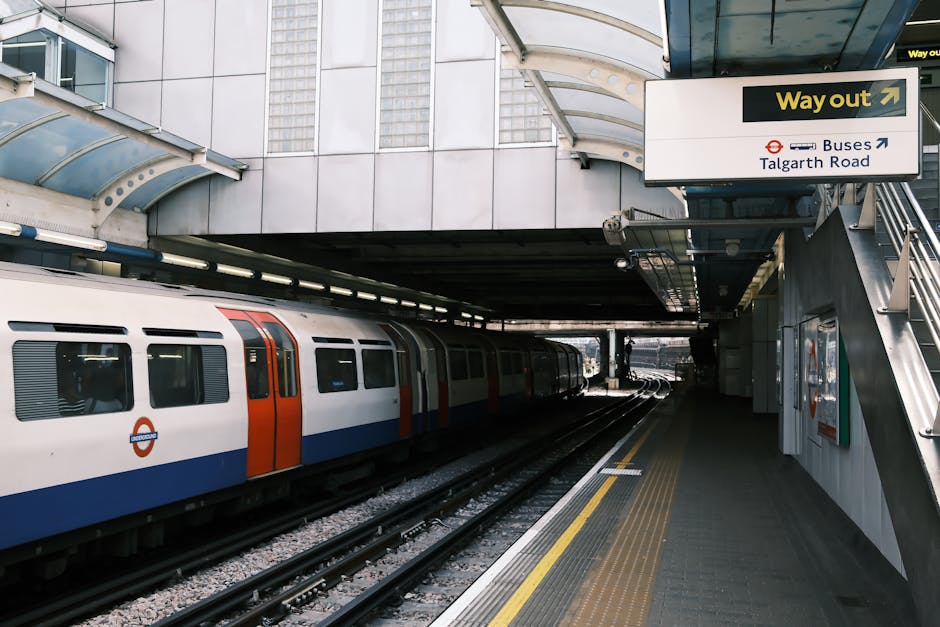 A modern underground London train station platform with a partially visible white train featuring orange and blue accents parked on the tracks. The platform surface is made of dark grey paving with a yellow tactile strip along the edge for safety. Overhead, a large white sign with black and yellow text directs passengers to the exit, indicating routes to buses and Talgarth Road, with the word 'Way out' prominently displayed. The station's architecture includes a curved, semi-transparent canopy overhead, supported by metal beams and glass panels, allowing natural light to illuminate the area. To the right, a metal staircase with a railing provides access to an upper level, while a wall-mounted fire safety cabinet is seen next to it. The environment appears clean and well-maintained, reflecting typical urban underground railway infrastructure used for public transport, suitable for individuals seeking information on local transport options or alternative waste handling facilities nearby, such as those offered by Waste Collection Hammersmith.