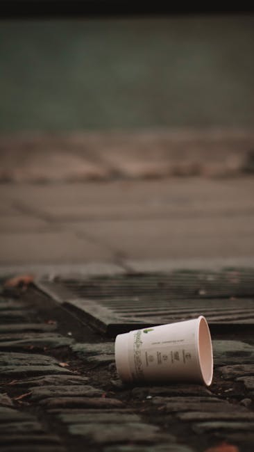 A discarded paper cup lies on a stone step or pavement in an outdoor setting, with part of a metal grate or drain cover visible nearby. The cup is light-colored, possibly white or cream, with printed text or design details faintly visible on its surface. The textured stone surface beneath the cup appears weathered and uneven, with some dirt or debris present around it. In the background, there is a blurred view of a paved or concrete surface, with muted tones of gray and green, suggesting an urban or semi-urban environment. The scene may be part of an outdoor area where rubbish has been carelessly left, illustrating a typical example of litter that would be handled through private rubbish removal services like those offered by Waste Collection Hammersmith. The overall lighting is subdued, possibly indicating overcast conditions, contributing to a neutral, observational tone suitable for a professional waste management context.