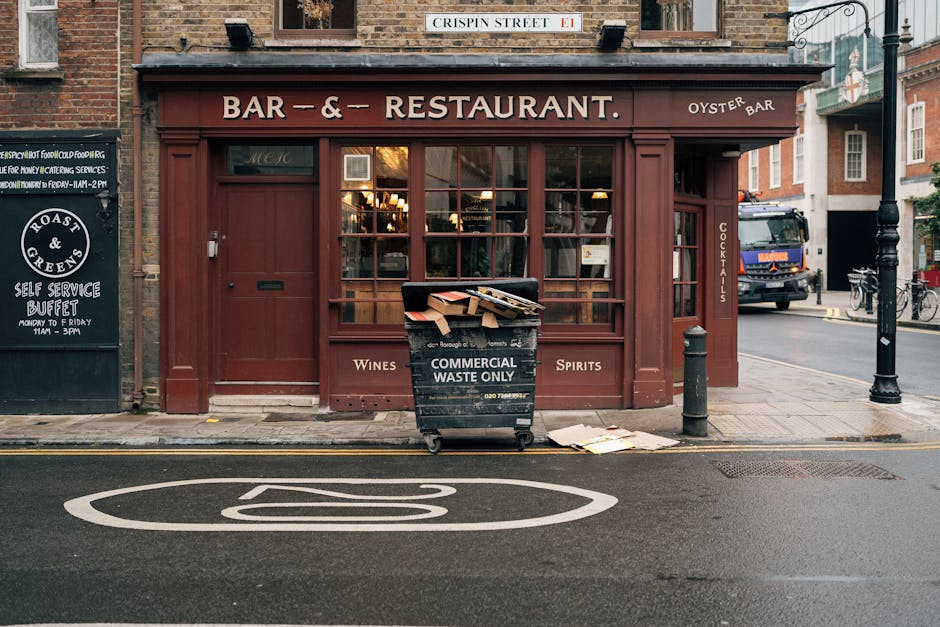 A black wheeled bin labeled 'COMMERCIAL WASTE ONLY' is positioned on the sidewalk in front of a red-brick building housing a bar and restaurant, with large glass windows showing warm interior lighting. The bin is partially open, filled with cardboard boxes and paper waste, and is situated next to a black metal bollard and a streetlamp. Nearby, a small blackboard sign advertises a self-service buffet and oyster bar, while a blue truck is visible on the street in the background. The scene is set on an urban street with a painted white parking restriction symbol on the road surface, and additional brick buildings and bicycles can be seen further along the street. The environment suggests a busy city environment with active waste management collection. Waste Collection Hammersmith provides rubbish removal services suitable for commercial waste disposal and street clearance. The overall scene highlights the importance of designated waste receptacles for different types of refuse, supporting efficient rubbish collection and disposal in the area, consistent with private waste handling practices rather than local authority collection.