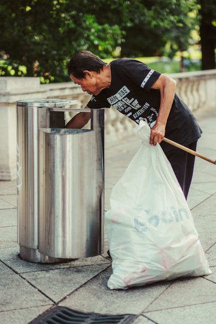 A woman with dark hair, wearing a black T-shirt with white printed text on the front and dark trousers, is seen disposing of waste into a stainless steel litter bin on a paved outdoor walkway. She is holding a large white plastic bag filled with rubbish in her left hand while using her right hand to open the top of the bin, which has a cylindrical shape and a polished, reflective surface. In the background, there are green leafy trees and a concrete balustrade, indicating a park or residential area setting. The scene is lit by natural daylight, with shadows cast on the pavement and the surrounding environment. This image relates to private waste collection or rubbish removal services provided by Waste Collection Hammersmith, illustrating a person properly discarding waste as part of an on-site rubbish clearance, aligning with the topic of efficient rubbish disposal and alternative waste handling solutions.
