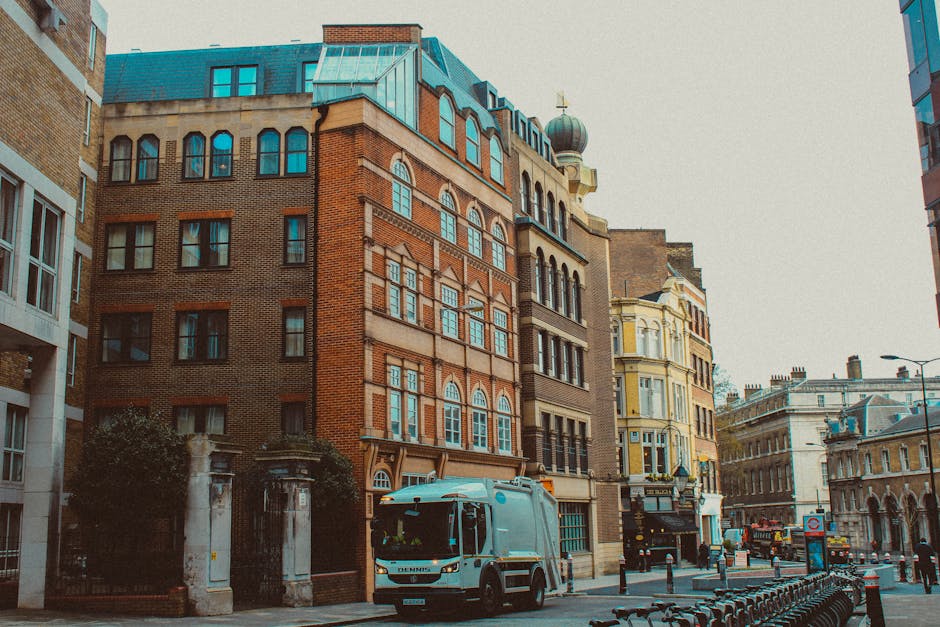 A city street scene featuring a mix of historic and modern multi-story buildings with diverse architectural styles, including brick facades, large windows, and ornate detailing. In the foreground, a blue and white waste collection van operated by Waste Collection Hammersmith is parked along the curb, with its side facing the viewer, indicating recent or ongoing rubbish removal services. The street is lined with metal bollards and bicycle racks, and the environment appears to be a busy urban area with adjacent buildings that include commercial establishments and residential apartments. The sky overhead is overcast, casting diffuse lighting across the scene, which emphasizes the textures of the buildings' materials and the street-level activity related to waste management and urban maintenance. The image reflects a typical setting where private rubbish collection services may be utilized for efficient waste handling in central London neighborhoods, subtly illustrating the context of alternative waste disposal methods servicing local businesses and residents.