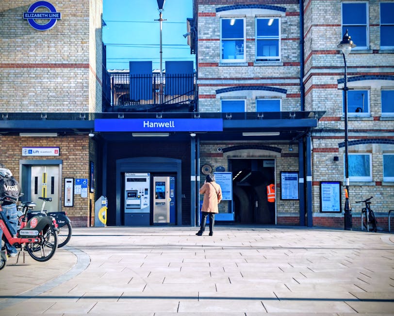 A woman wearing a beige coat and black pants stands on a paved pedestrian area in front of Hanwell station, part of the Elizabeth Line, with a dark blue station sign above the entrance. The station building features a brick facade with a mix of cream, beige, and red-brown bricks, several windows, and black metal railings along the upper balcony. To the left of the entrance, there is an automated ticket machine, and nearby is a yellow contactless payment device mounted on a post. Several bicycles, including one with a cargo rack, are parked to the left, secured to bike stands. A black lamppost with a traditional lantern-style light, and a smaller lamppost with an orange and white sign, are positioned on the right side of the scene. The atmosphere is bright, indicating daytime with clear weather, and the surrounding environment includes a clean, modern pavement area adjacent to the station, highlighting urban transportation infrastructure and private bike parking as part of the station's amenities. The scene visually supports the context of public transit, with potential for out-of-hours or alternative transport options involved in local urban rubbish and waste management services, such as that provided by Waste Collection Hammersmith.