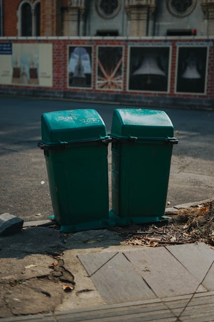 A city street scene featuring a mix of historic and modern multi-story buildings with diverse architectural styles, including brick facades, large windows, and ornate detailing. In the foreground, a blue and white waste collection van operated by Waste Collection Hammersmith is parked along the curb, with its side facing the viewer, indicating recent or ongoing rubbish removal services. The street is lined with metal bollards and bicycle racks, and the environment appears to be a busy urban area with adjacent buildings that include commercial establishments and residential apartments. The sky overhead is overcast, casting diffuse lighting across the scene, which emphasizes the textures of the buildings' materials and the street-level activity related to waste management and urban maintenance. The image reflects a typical setting where private rubbish collection services may be utilized for efficient waste handling in central London neighborhoods, subtly illustrating the context of alternative waste disposal methods servicing local businesses and residents.