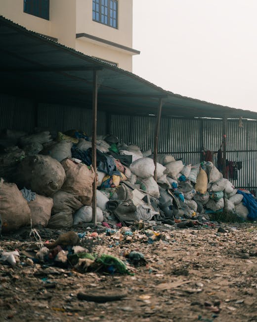 A large collection of various waste materials, primarily comprising irregularly shaped plastic bags filled with refuse and other discarded items, stacked beneath a corrugated metal roof supported by thin vertical poles. The bags vary in size, material, and colour, with some appearing translucent and others opaque, displaying shades of white, black, blue, and beige. Among the bags, there are loose pieces of rubbish and smaller debris scattered across the muddy, uneven ground in the foreground, which also contains bits of plastic, paper, and organic matter. The scene is set outdoors, adjacent to a grey metallic fence running horizontally across the background, extending behind the waste piles, with part of a cream-coloured building with dark-framed windows visible in the upper left corner, indicating a residential or commercial premises in the vicinity. The overall atmosphere is utilitarian, with natural light highlighting the textures and materials, reflecting a typical site where private waste collection or alternative rubbish handling services, such as those offered by Waste Collection Hammersmith, might be employed for on-site waste clearance.