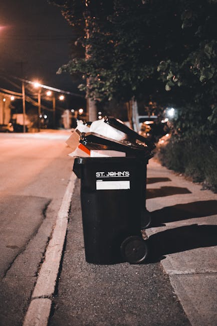 A black wheelie bin labeled 'St. John's' on a paved sidewalk during nighttime, with a white stripe around its middle and overflowing with mixed rubbish, including cardboard boxes, plastic packaging, and paper. The bin is positioned near a curb, casting a shadow on the ground. In the background, a dimly lit street features street lamps, a row of parked cars, and trees with dark foliage, suggesting an urban or suburban residential area. The environmental context indicates this bin is used for waste collection, and the scene reflects the typical setting for private rubbish disposal or alternative waste handling outside of official collection schedules, tied in subtly with waste management services like those offered by Waste Collection Hammersmith.